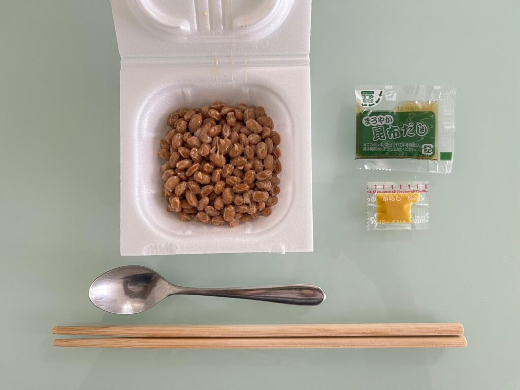 Opened pack of natto next to its sauce and mustard packets, with a spoon and chopsticks
