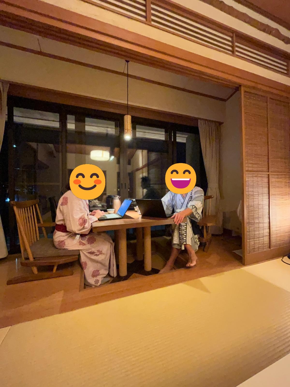 Two people in yukata working on laptops at a table in a ryokan room, faces covered with emoji